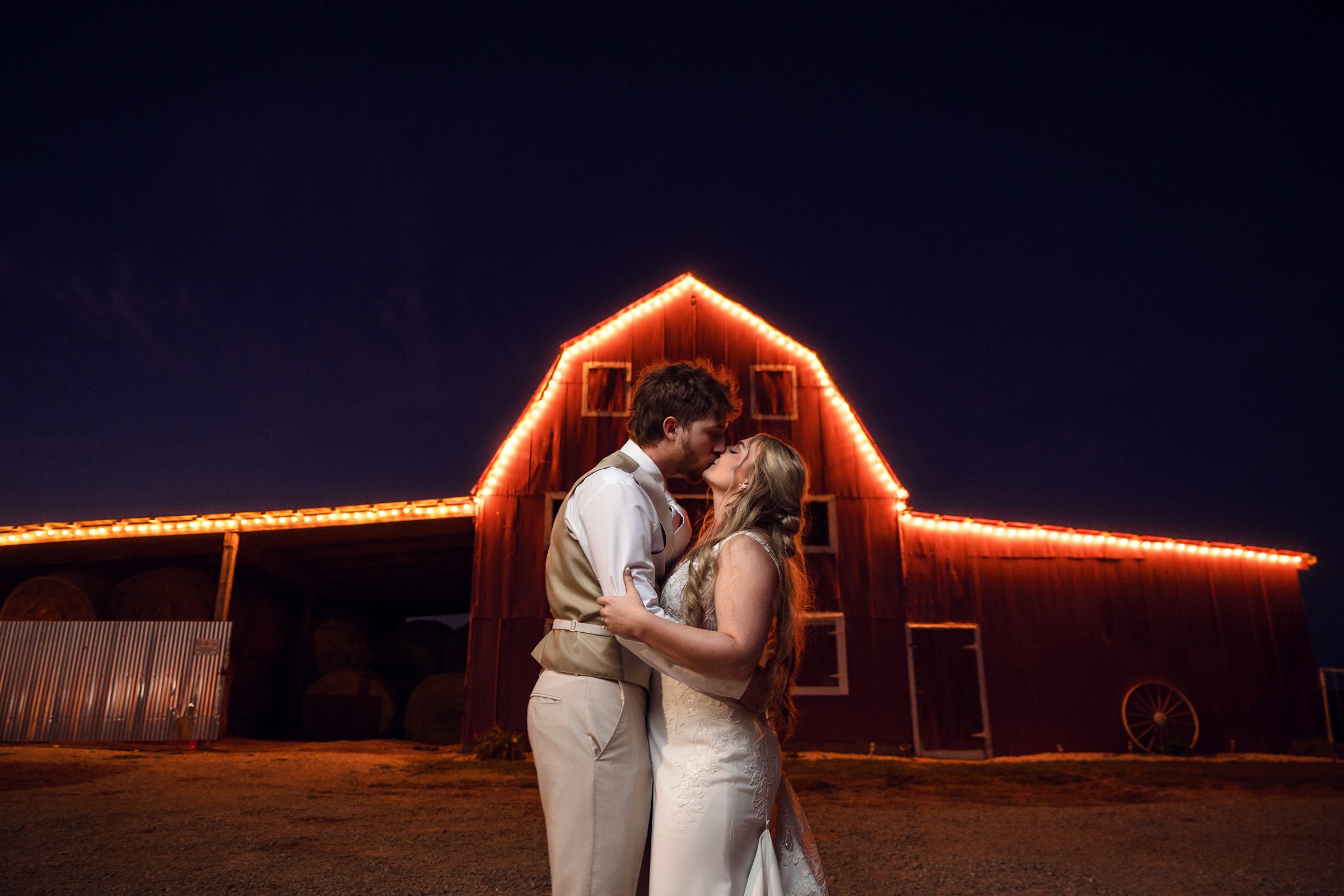 Bride and groom kiss in front of red barn on farm with christmas lights