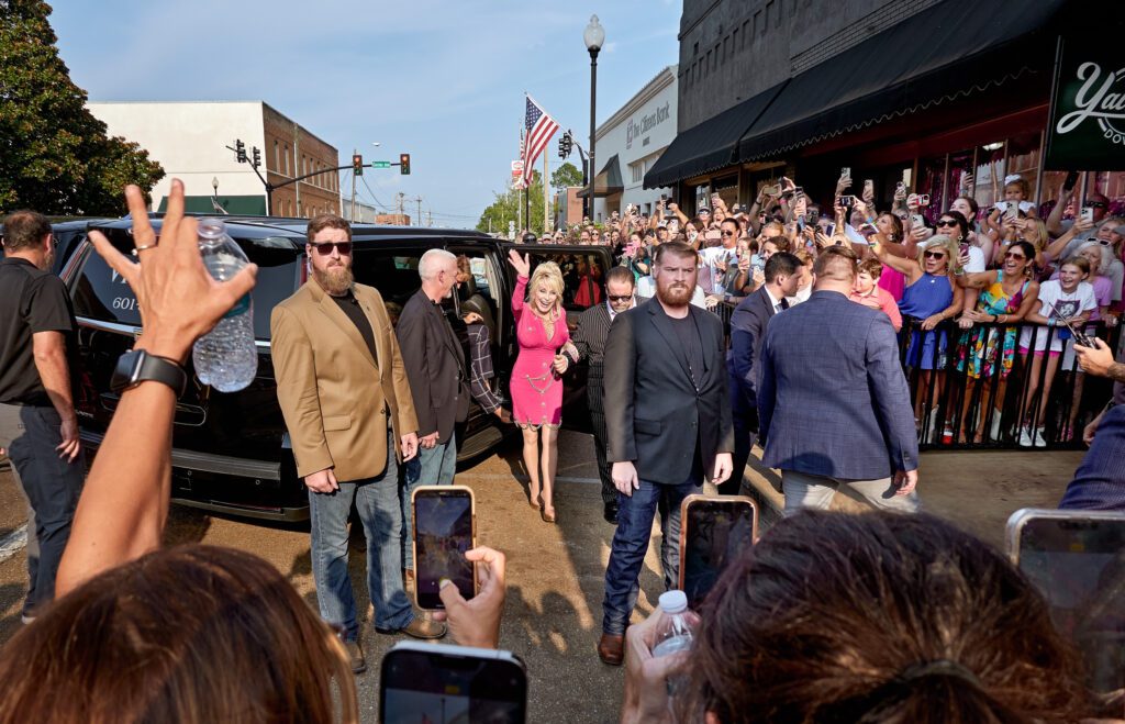 Dolly Parton waves at fans while security watches the crowd.