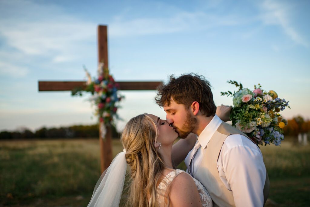 bride and groom kiss in front of cross with flowers in field