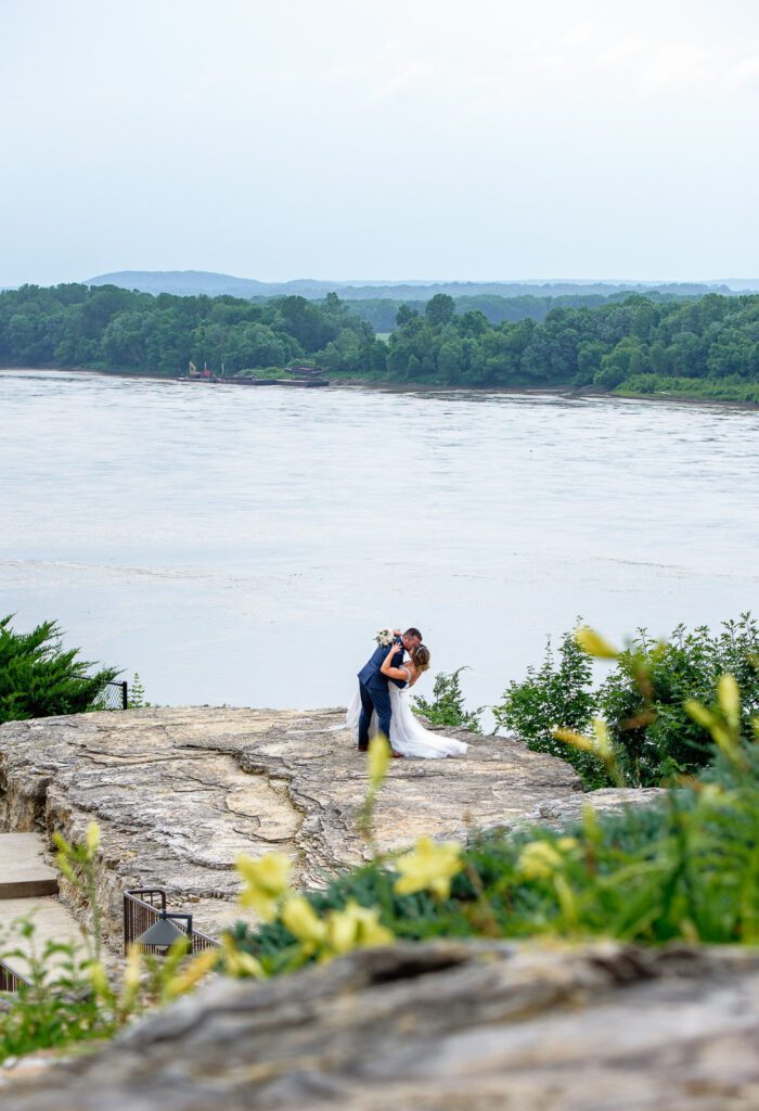 Bride and Groom kiss on cliff with river in the distance.