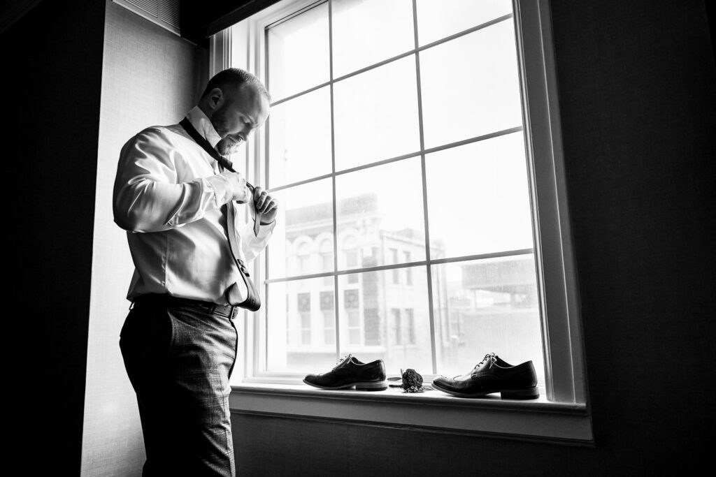 Groom adjusting tie in front of window