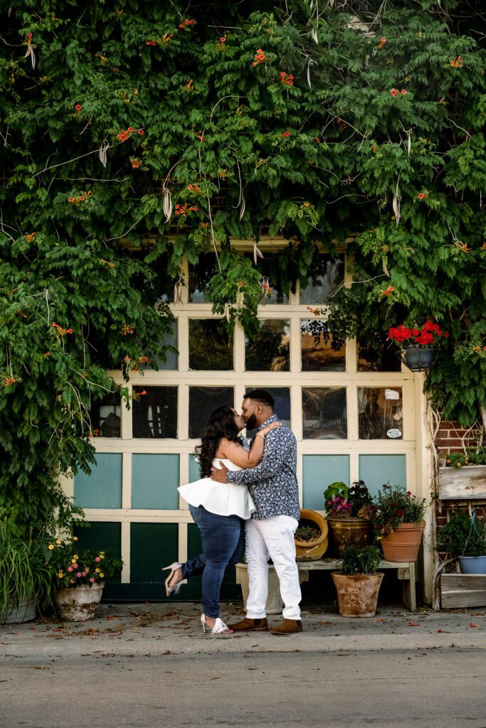 Engaged couple kiss in front of a garage door with green vines and red flowers