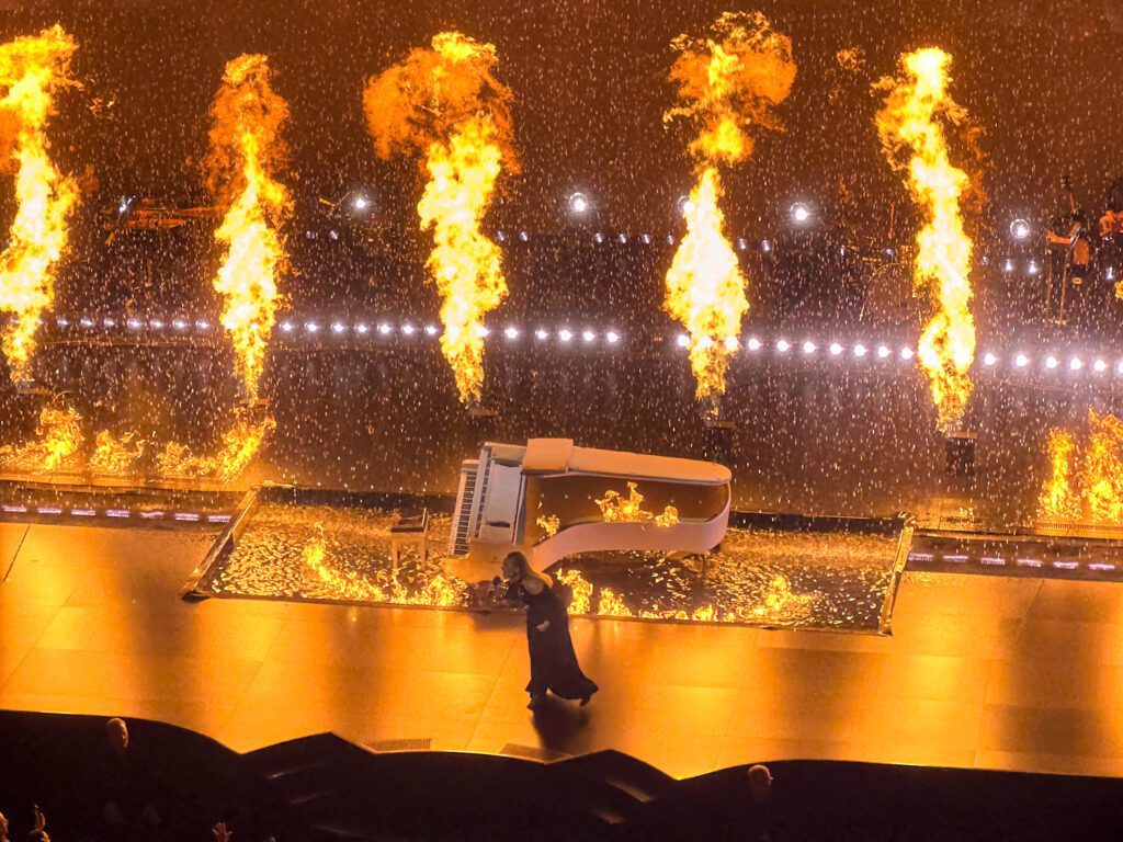 Adele sings fire and rain with fire columns shooting up in the back ground behind a burning piano.