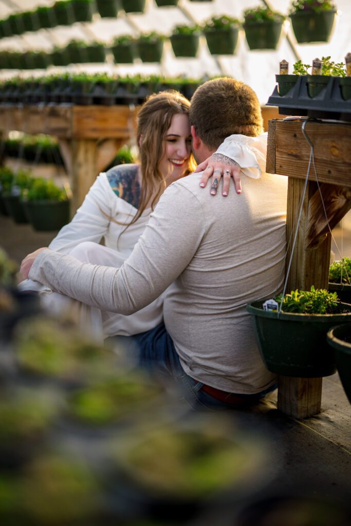 engaged couple laughing while sitting in green house with golden sunlight
