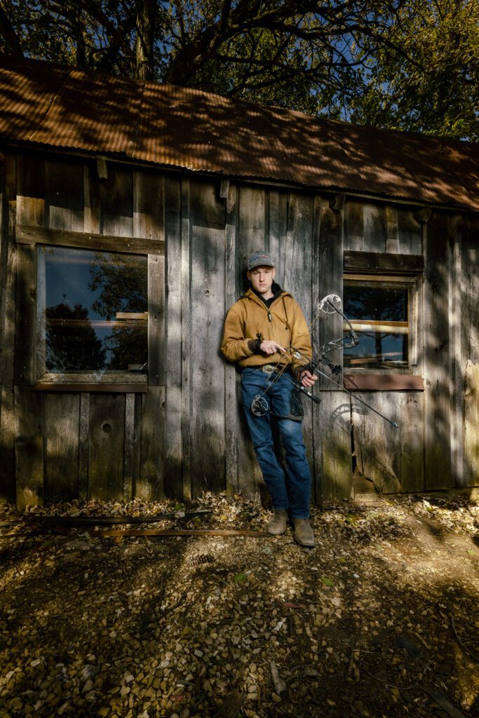 High school senior with bow and carhart jacket leans on old wood shed