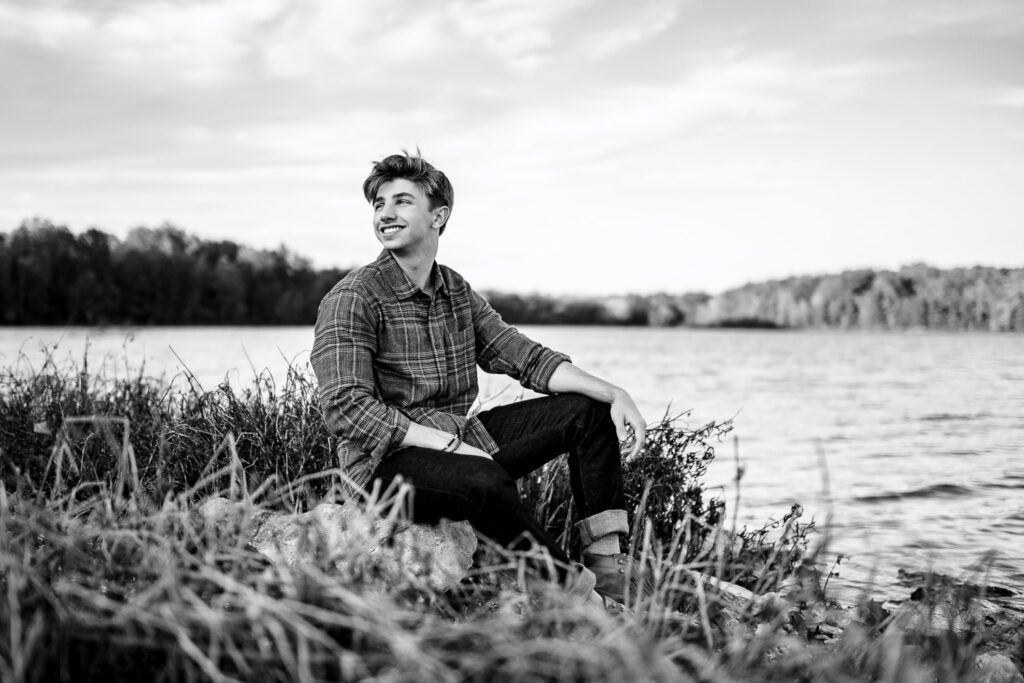 high school senior guy sitting by lake looking over his shoulder smiling