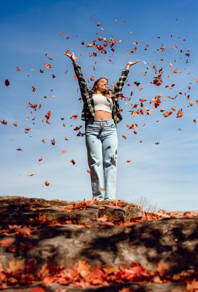 senior girl on top of rock cliff throws red and orange fall leaves into the air wearing white top and flannel