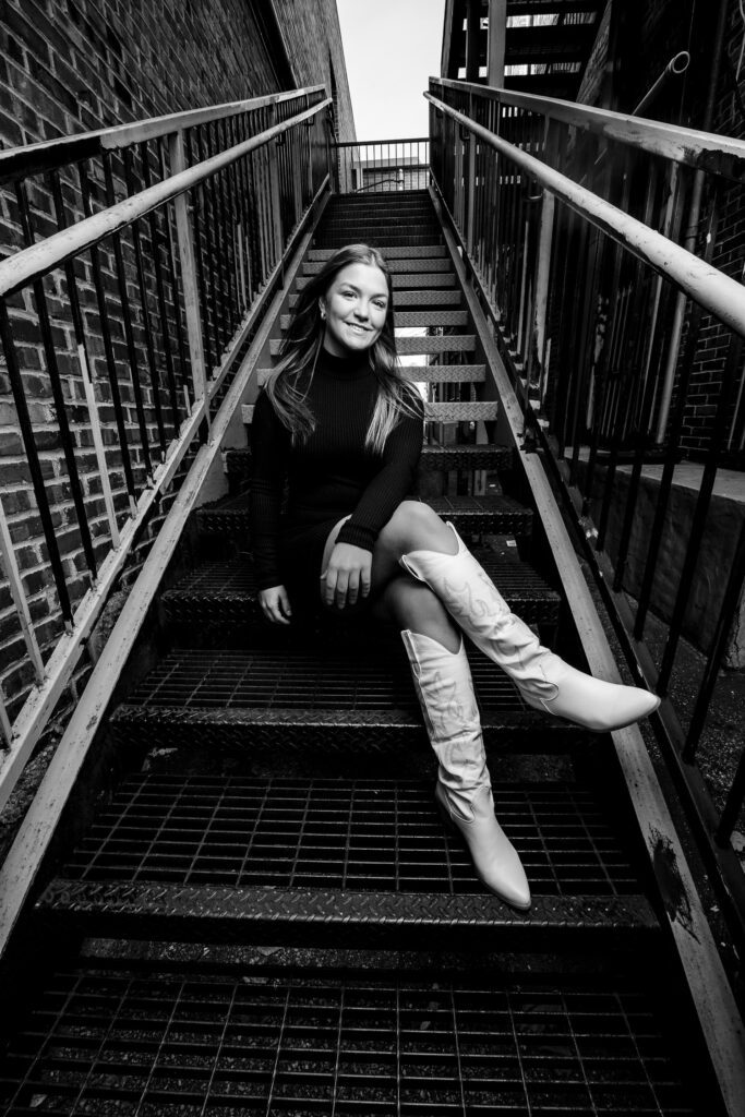 senior girl in columbia, missouri sits on metal staircase wearing black dress and white boots