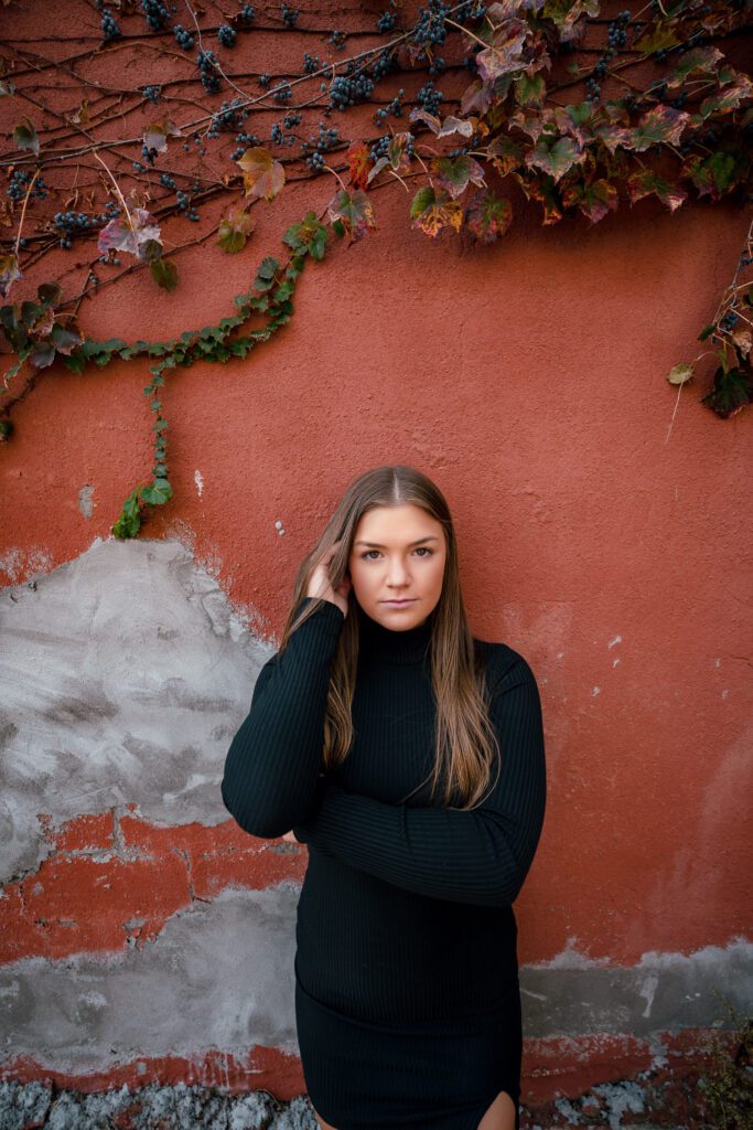 brunette senior high school girl leans on red and grey wall with green vines