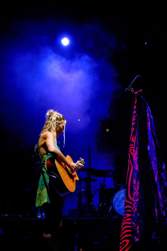 Lennon Stella playing acoustic guitar with purple scarf on mic.