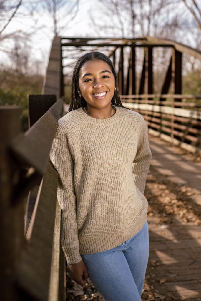 Black Senior girl wearing tan sweater stands on old railroad bridge at Capen Park