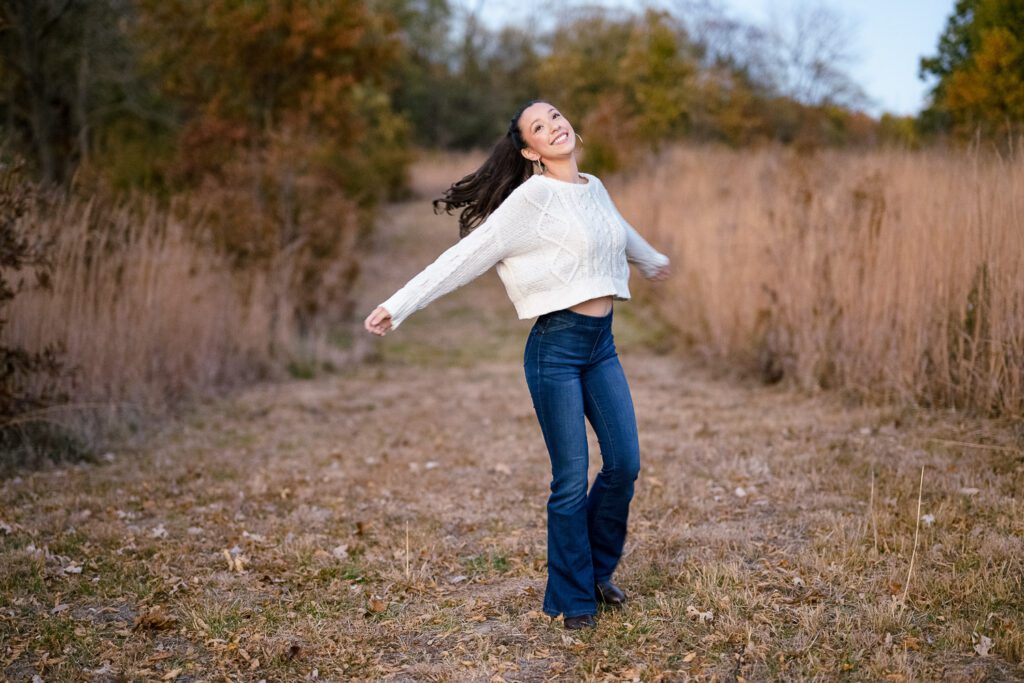 Senior girl wearing white sweater and blue jeans twirls in fall field near columbia, missouri.