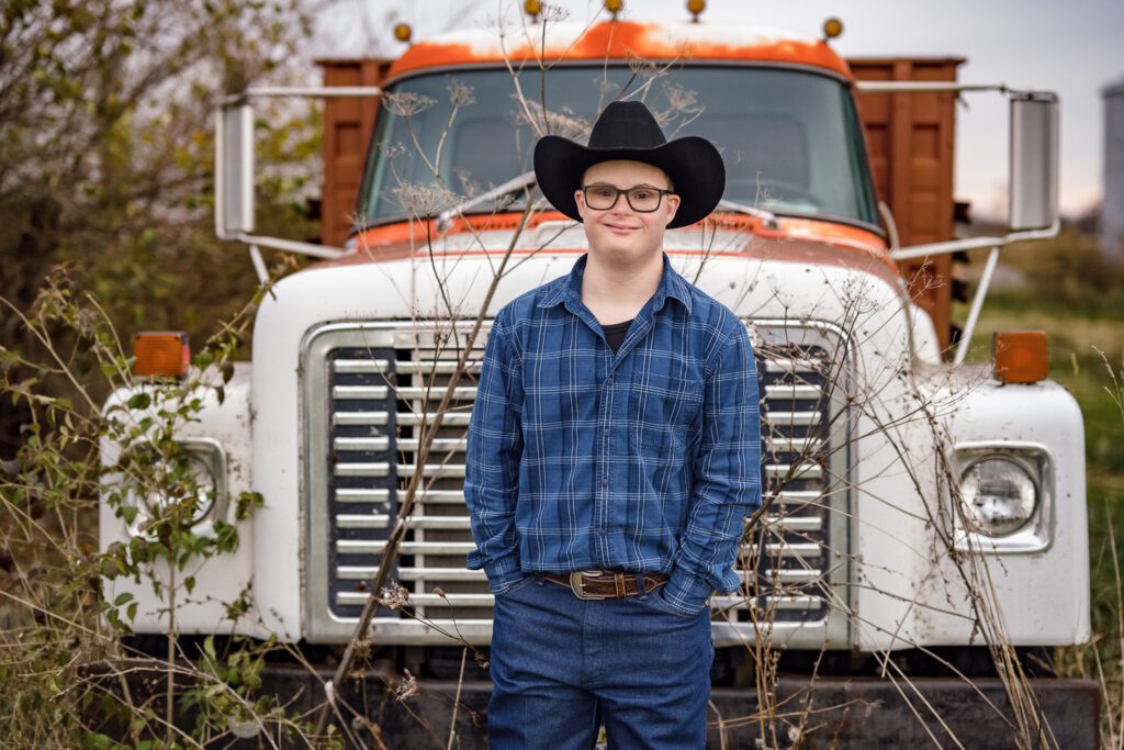 Senior guy with down syndrom wearing blue flannel shirt and black cowboy hat standing in front of old Mac Farm Truck