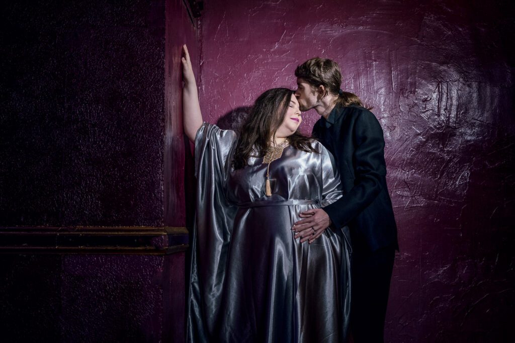 Expecting Mother leans on purple wall wearing silver maternity gown. Husband kisses her forehead.