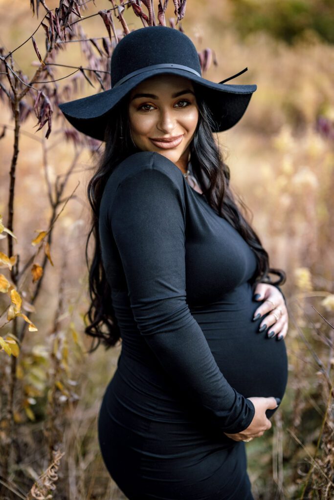Pregnant mother wearing black dress and hat holds pregnant belly during maternity shoot.