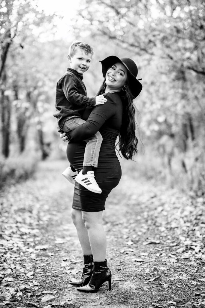 Pregnant mother wearing black dress holds son sitting on pregnant belly.