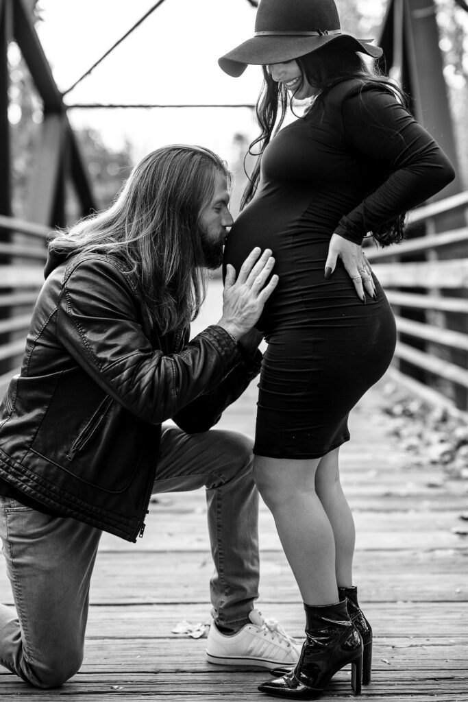 Guy kneels down to kiss pregnant belly of wife wearing black dress.