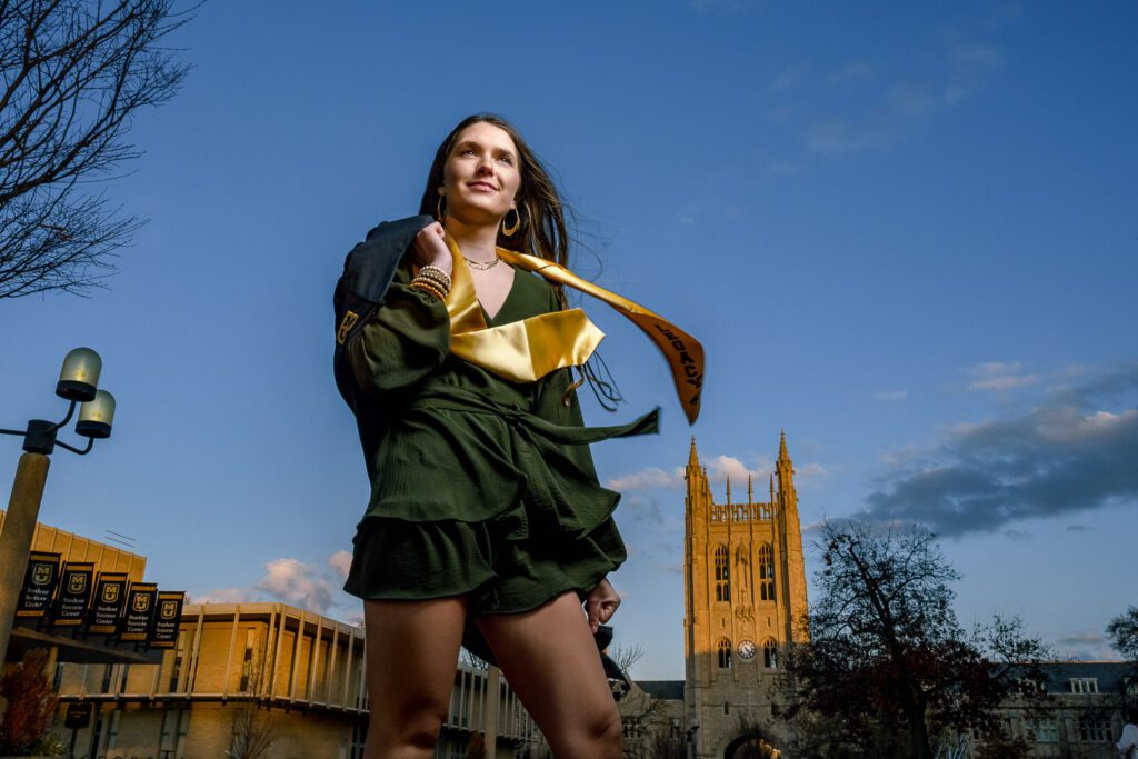 Senior girl wearing graduation gown stands in front of Mizzou's Memorial Union on a windy afternoon
