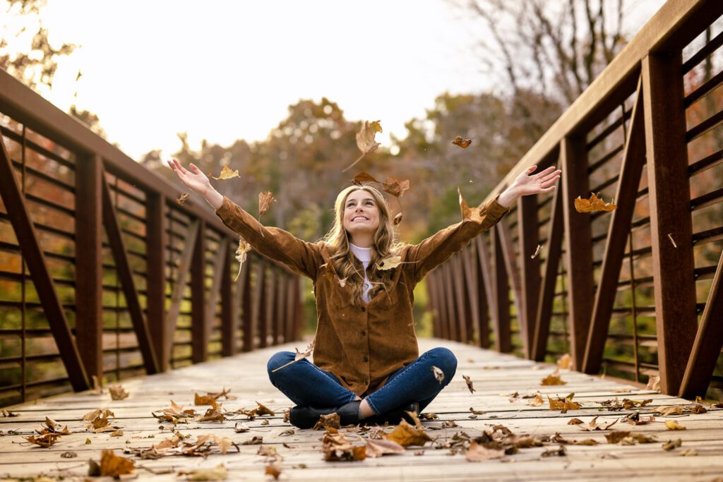 senior girl wearing brown top sits on bridge while throwing a handful of fall leaves in the air near columbia, missouri