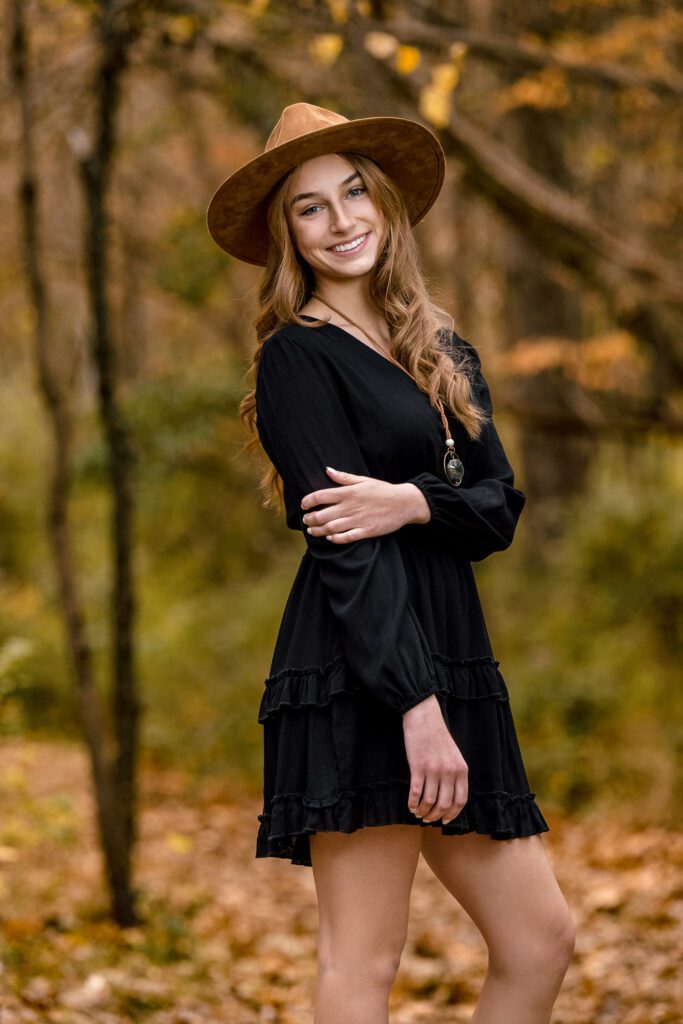 senior girl wearing brown hat and black dress holding arm in woods