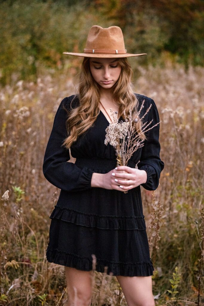 senior girl wearing brown hat and black dress holding fall grass and flowers in field