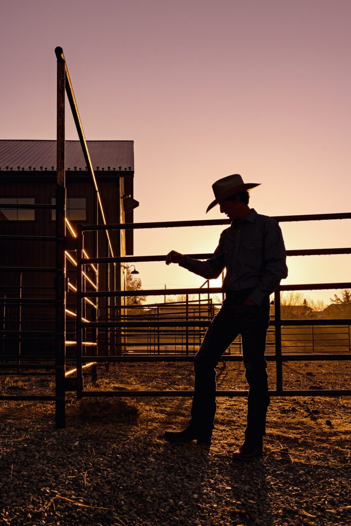 silhouette of high school senior wearing cowboy hat leaning on fence outside barn