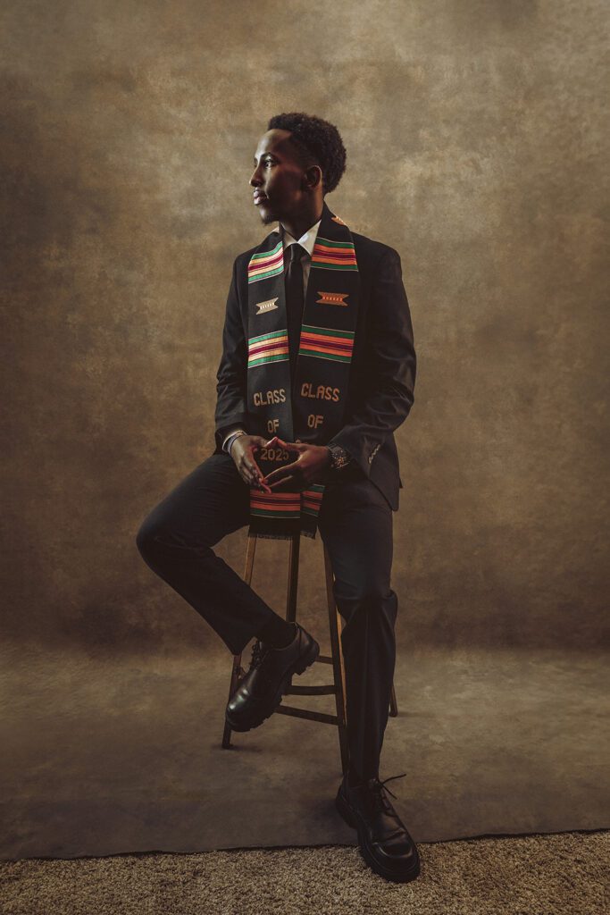 College Black Senior Guy sitting on stool with Kente Stoll and Mortar Board