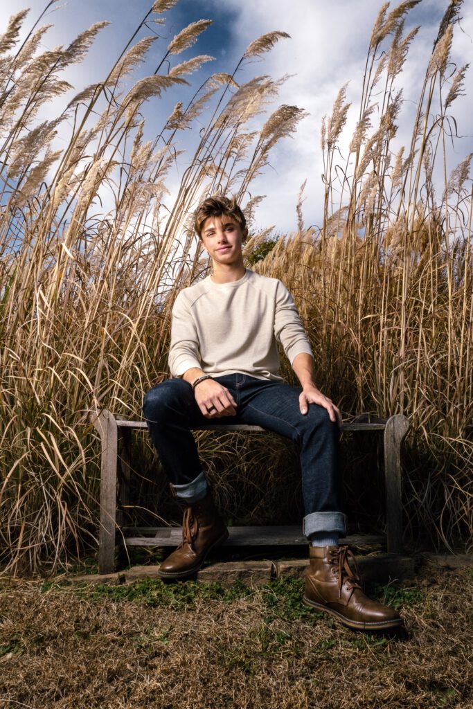 Senior guy with blue jeans and brown boots sits on bench in front of tall grass and clouds blowing in wind