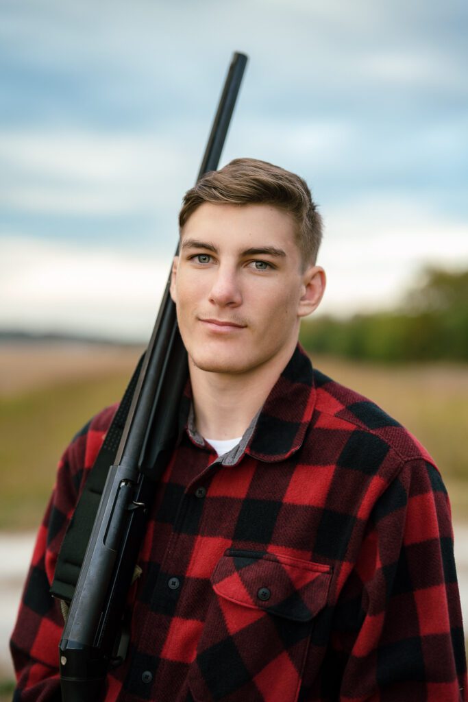 high school senior with buffalo plaid shirt and shotgun stands in field