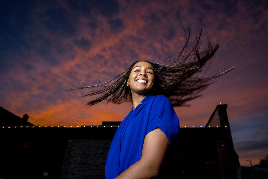 black senior girl wearing blue dress swings hair in air with beautiful pink and purple sky
