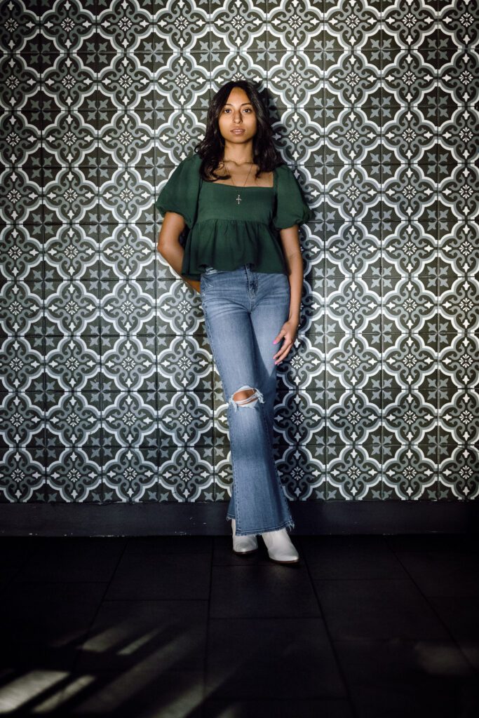 Senior girl wearing green top and blue jeans leans against ceramic tile patterned wall