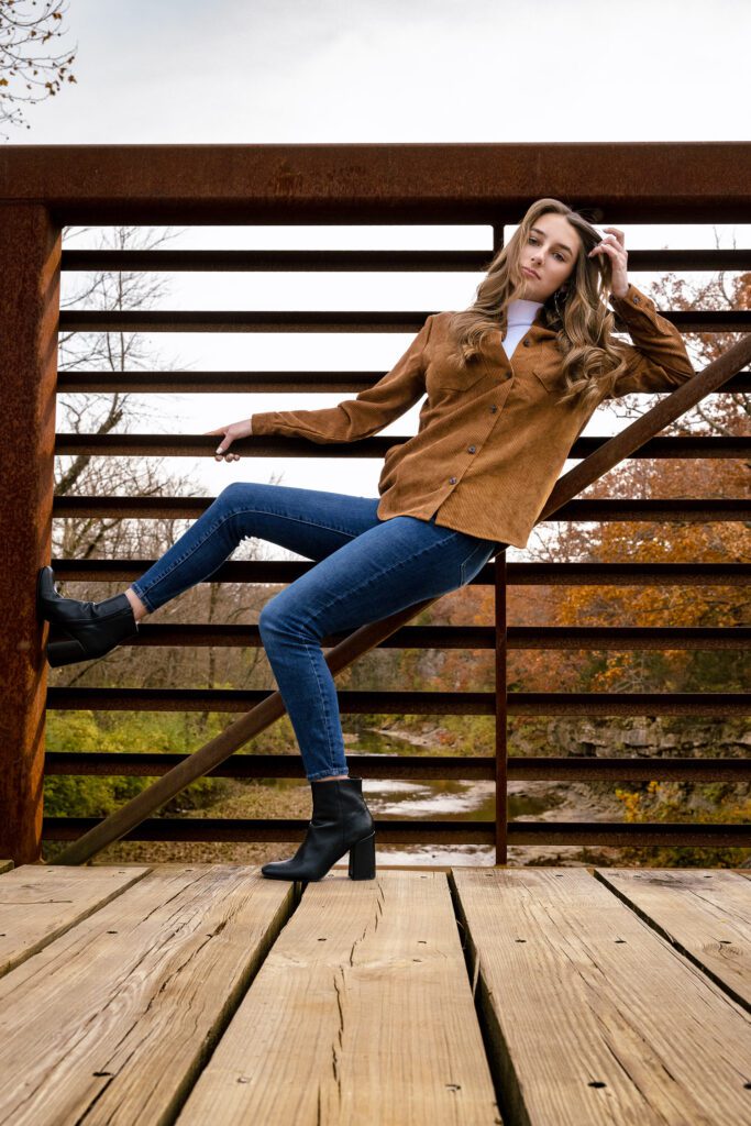senior girl with blue jeans and brown top lays on diagonal bridge railing