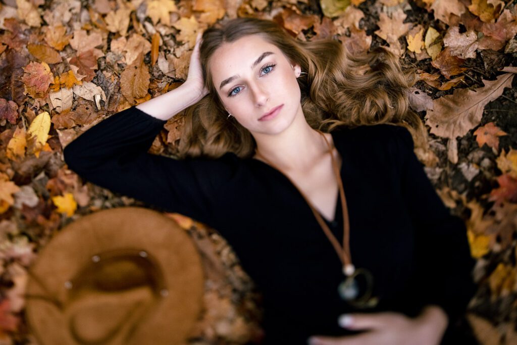 Senior girl wearing black dress lays on bed of fall leaves with brown hair and hat