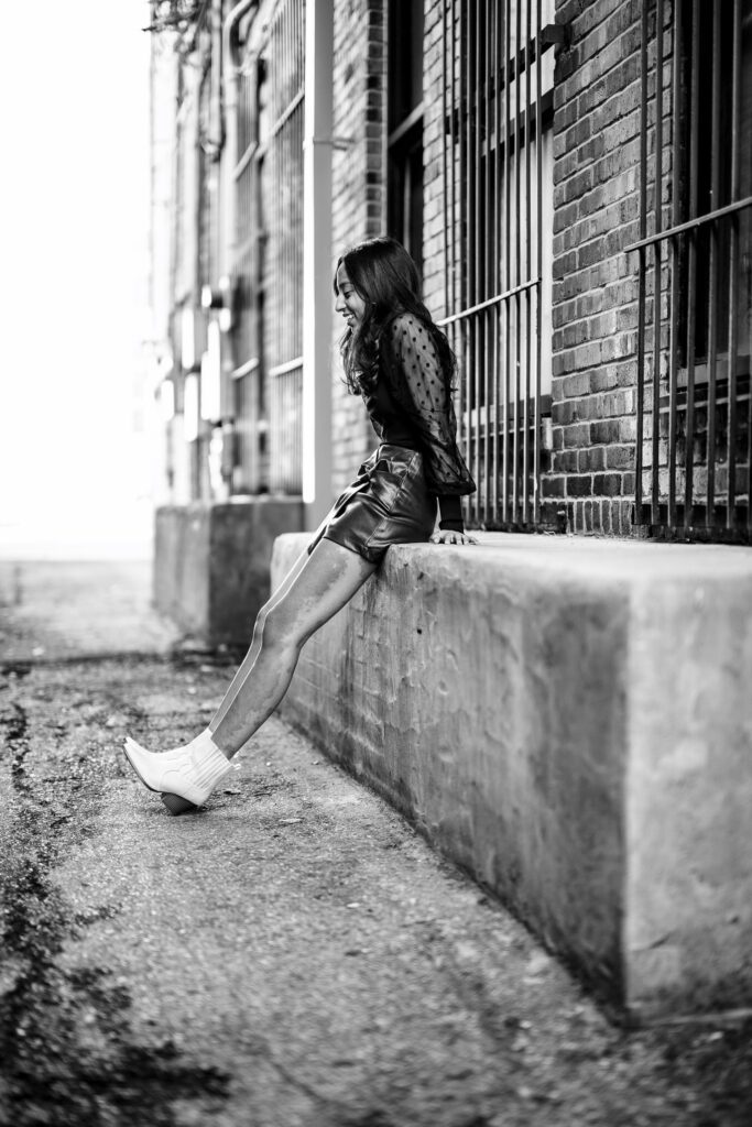 Senior girl wearing leather skirt and black top sits on concrete block in alley