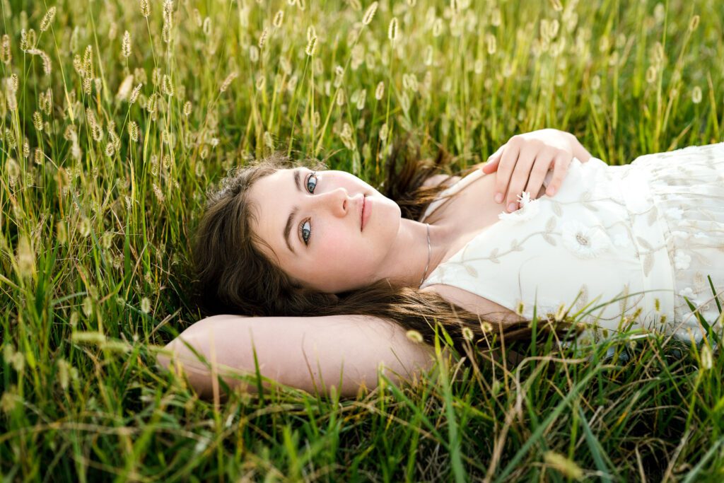 senior girl in white lace dress lays in grass field with one hand on her chest.
