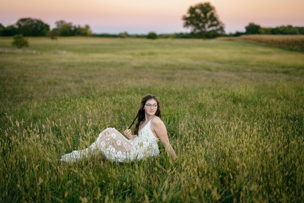 Senior girl in white lace dress sits in grass field at sunset.