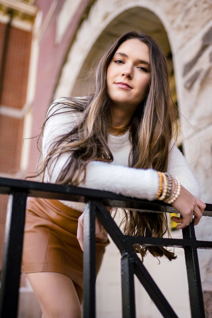 Senior girl wearing brown leather skirt and white top leans over black metal staircase railing.