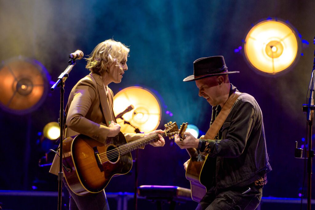 Brandi Carlile playing guitar with band under colorful stage lights.
