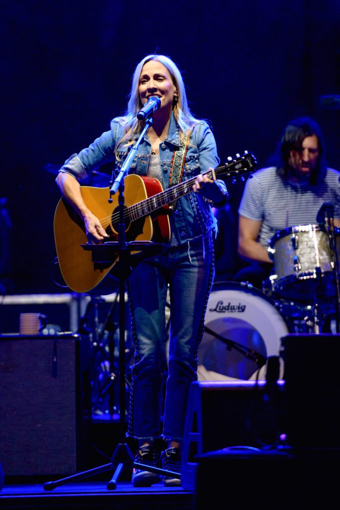 Sheryl crow sings while playing guitar on stage.