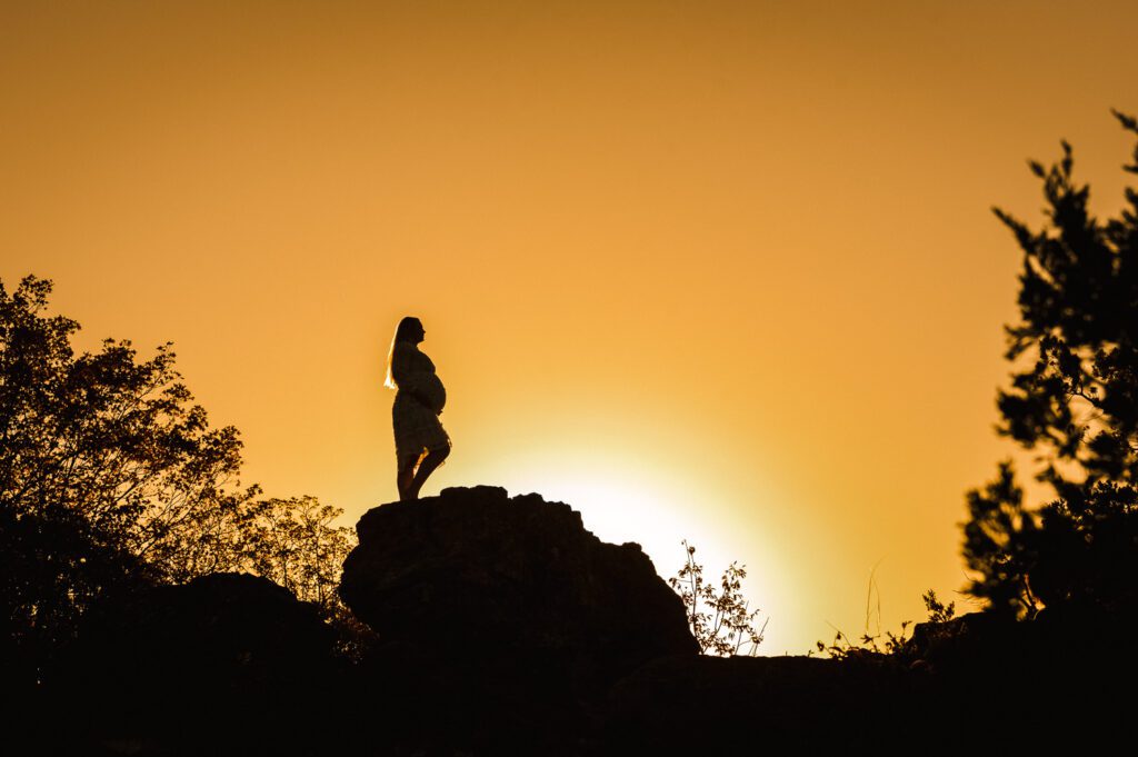 girl standing on giant rock holding pregnant belly at sunset