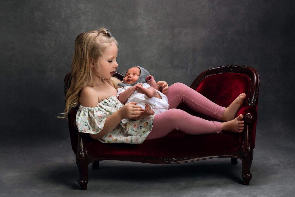 sister holding newborn son with bonnet on tiny red couch