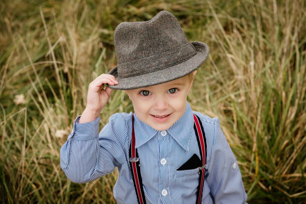little boy tipping fedora hat in field in suspenders