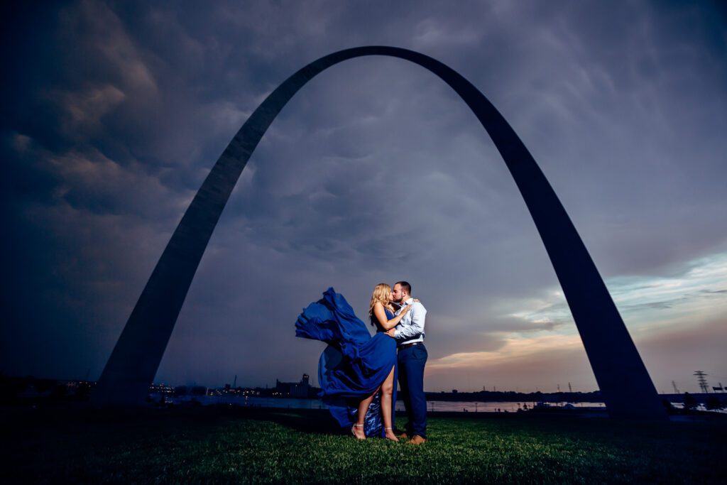 Engaged couple kissing under St. Louis Arch with dress flying