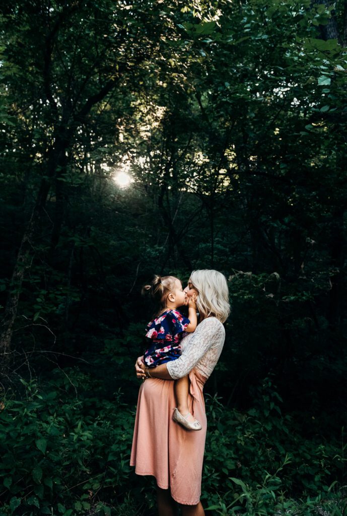 pregnant mother holding daughter on stomach kissing in woods