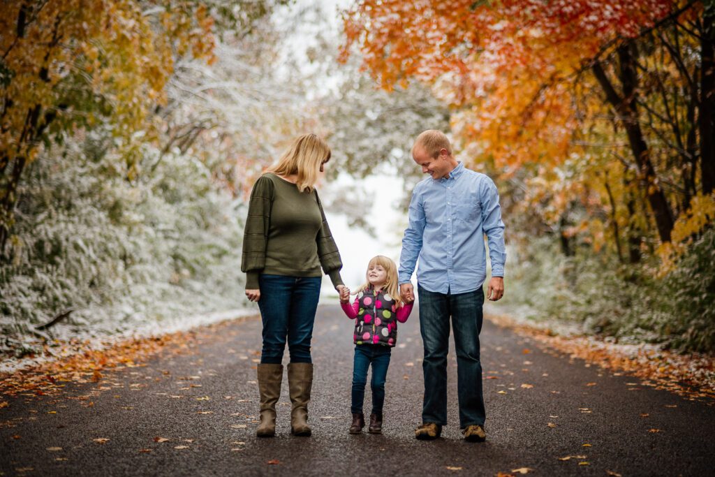 mother and father holding daughters hand in snowy roadway with fall leaves