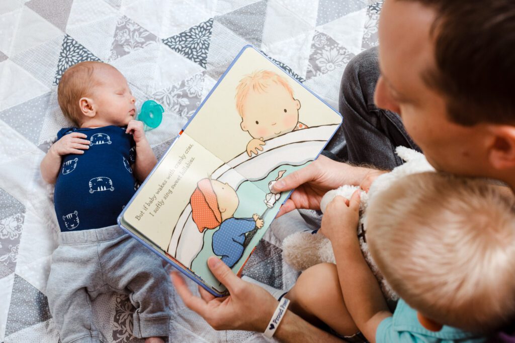 father reading book to son and newborn baby on quilt
