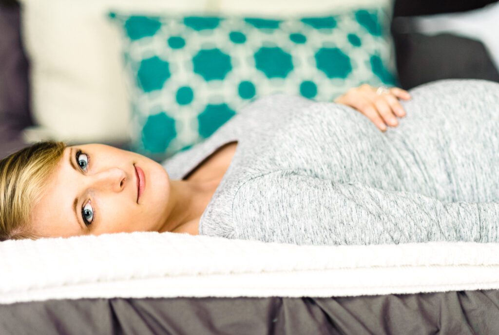 woman laying on bed looking in camera while holding pregnant belly