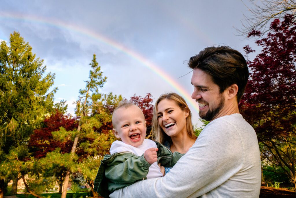 A family of three smile under rainbow.