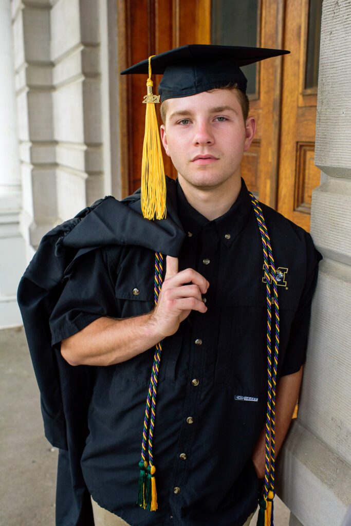 Senior guy holds gown over shoulder while wearing cap.