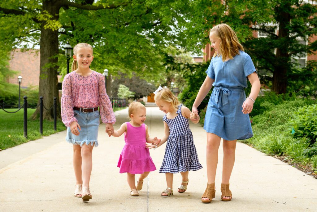Four sisters hold hands while walking on sidewalk.